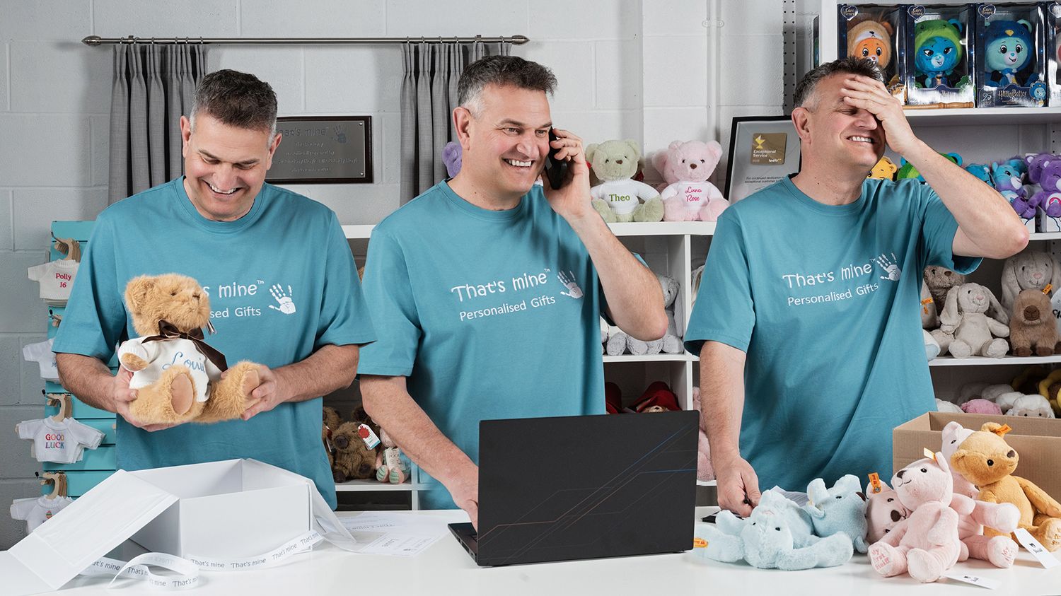 Three men in teal shirts smile and laugh around a table filled with colorful stuffed animals and toys.