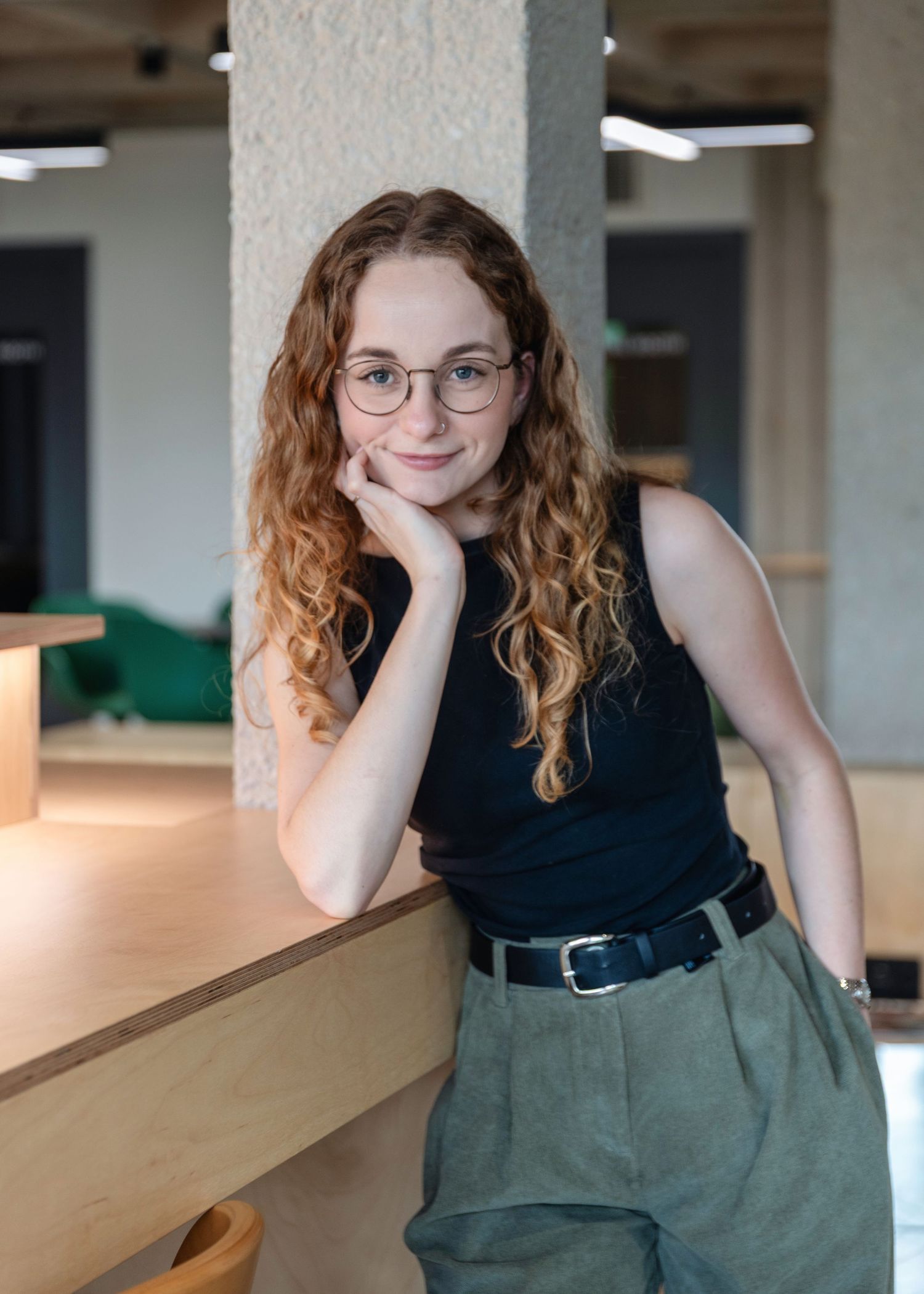 Young woman with curly red hair and glasses poses casually in a modern office space wearing a black top and olive pants.