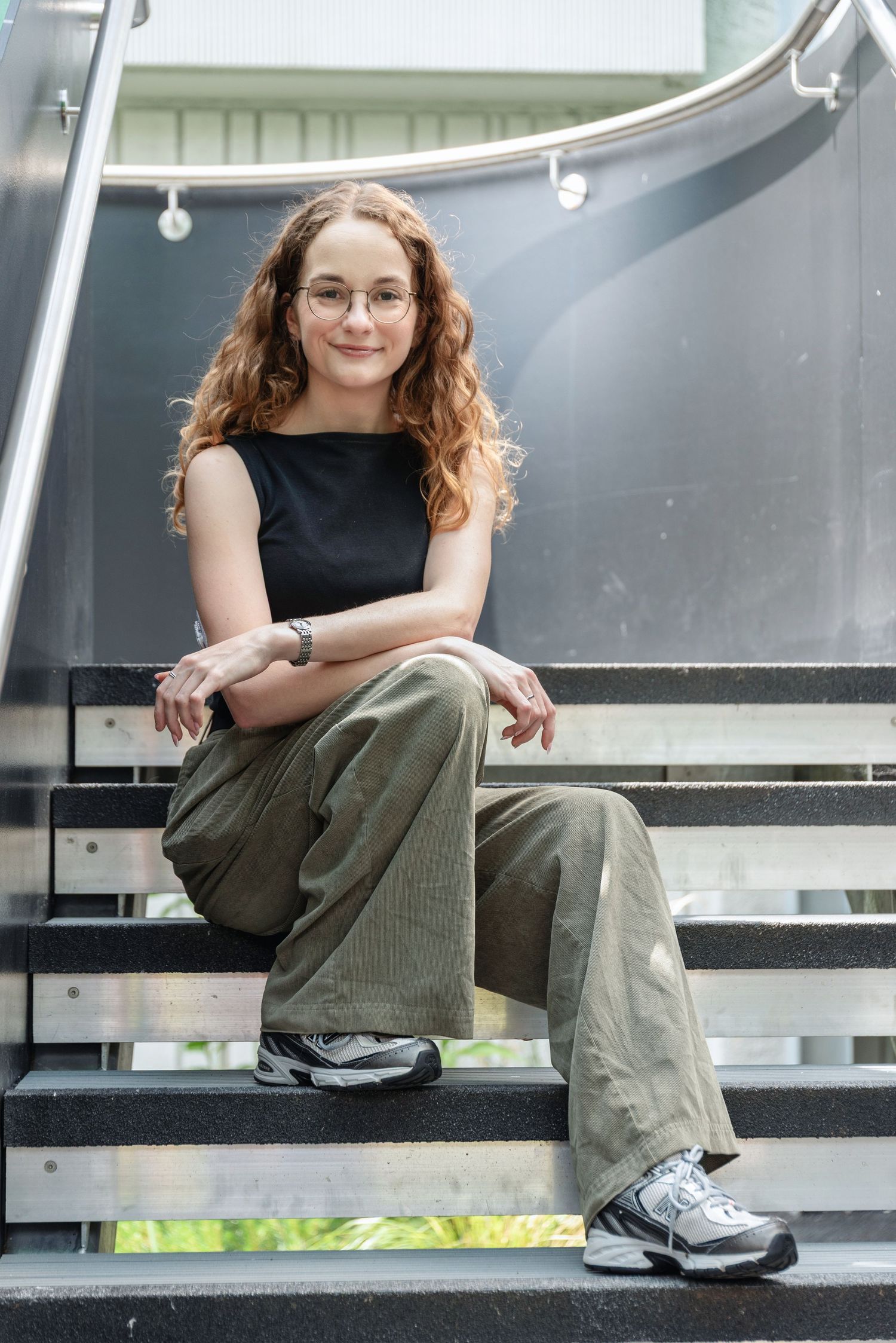 Young woman with curly red hair and glasses sits relaxed on modern stairs wearing olive cargo pants and black top.