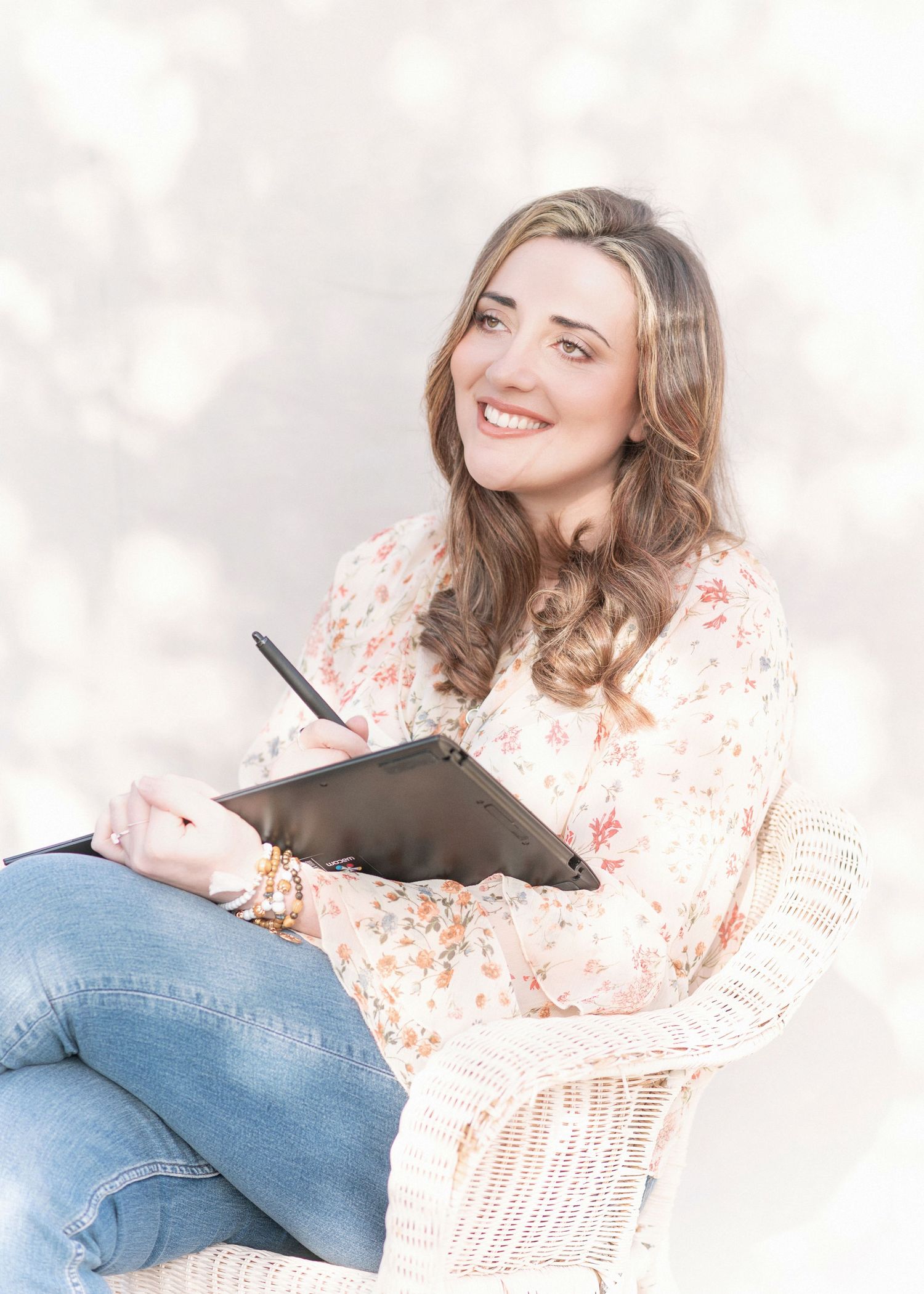 Smiling woman in floral blouse and jeans sits in white wicker chair, writing in a notebook against a soft bokeh background.