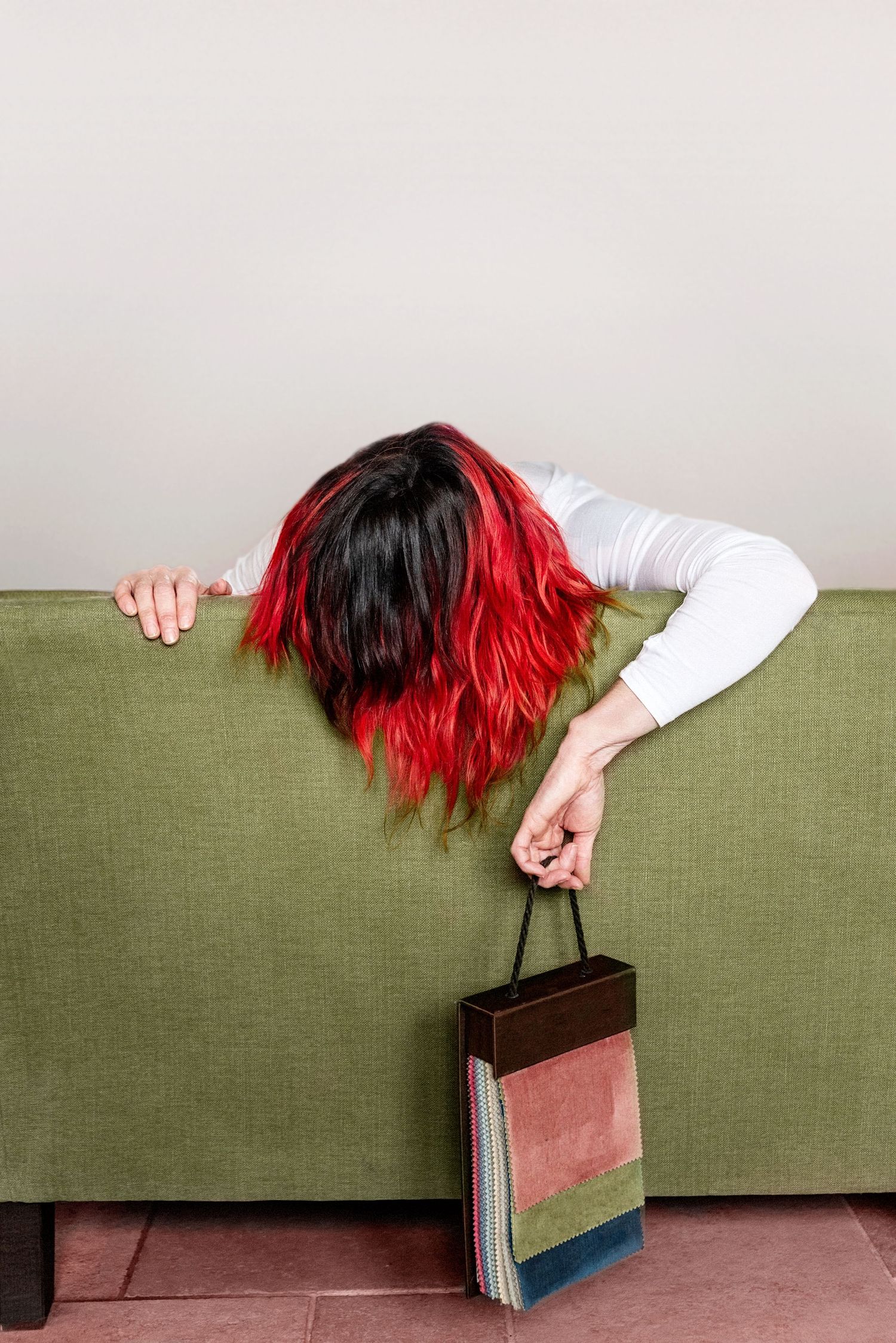 Person with red-dipped black hair draped over green couch holding colorful books while appearing exhausted.
