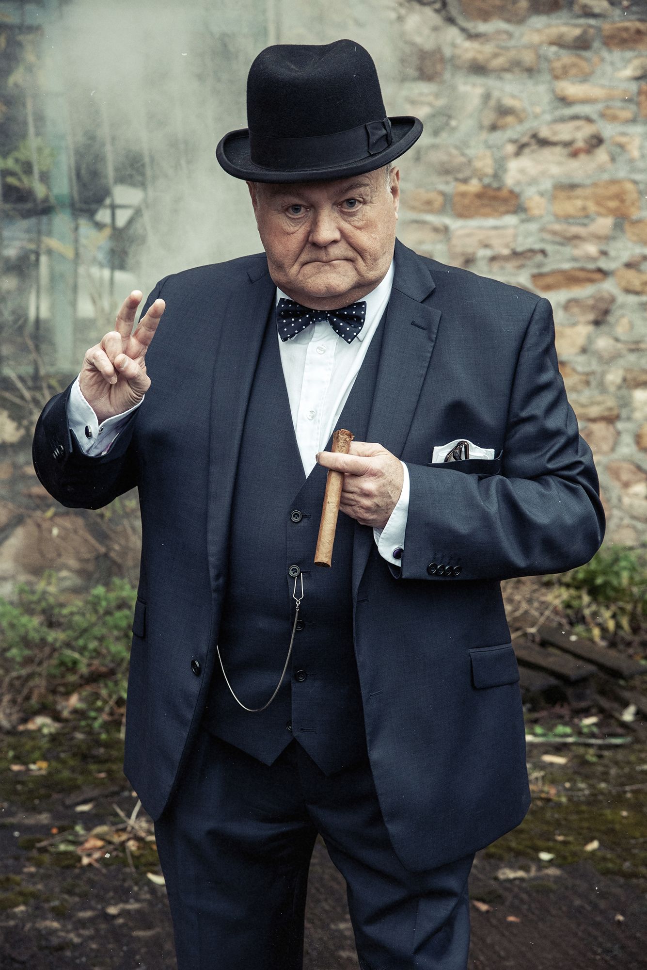 Gentleman in bowler hat and suit makes peace sign while posing against stone wall.