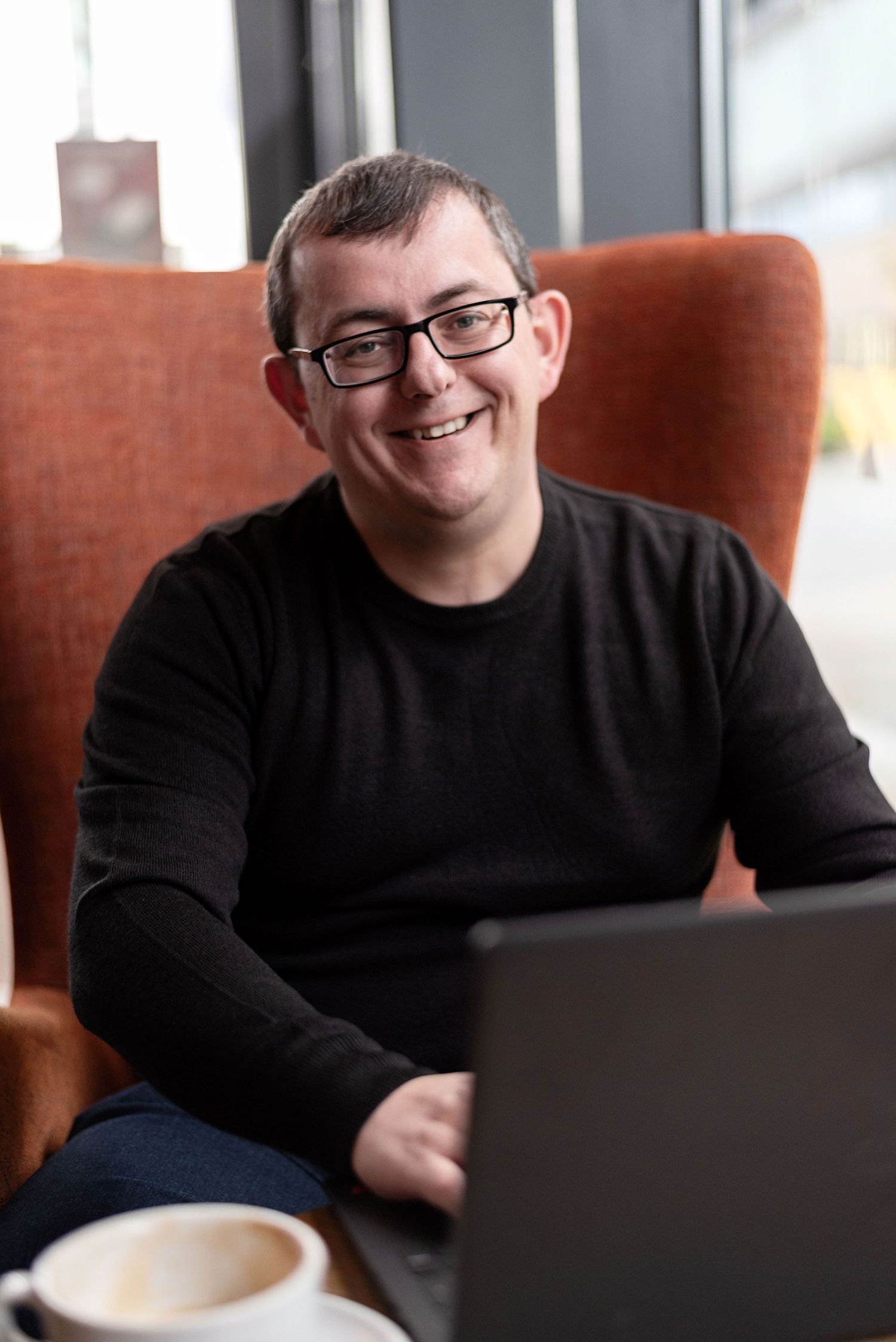 Person in black sweater working on laptop in orange armchair with coffee cup.