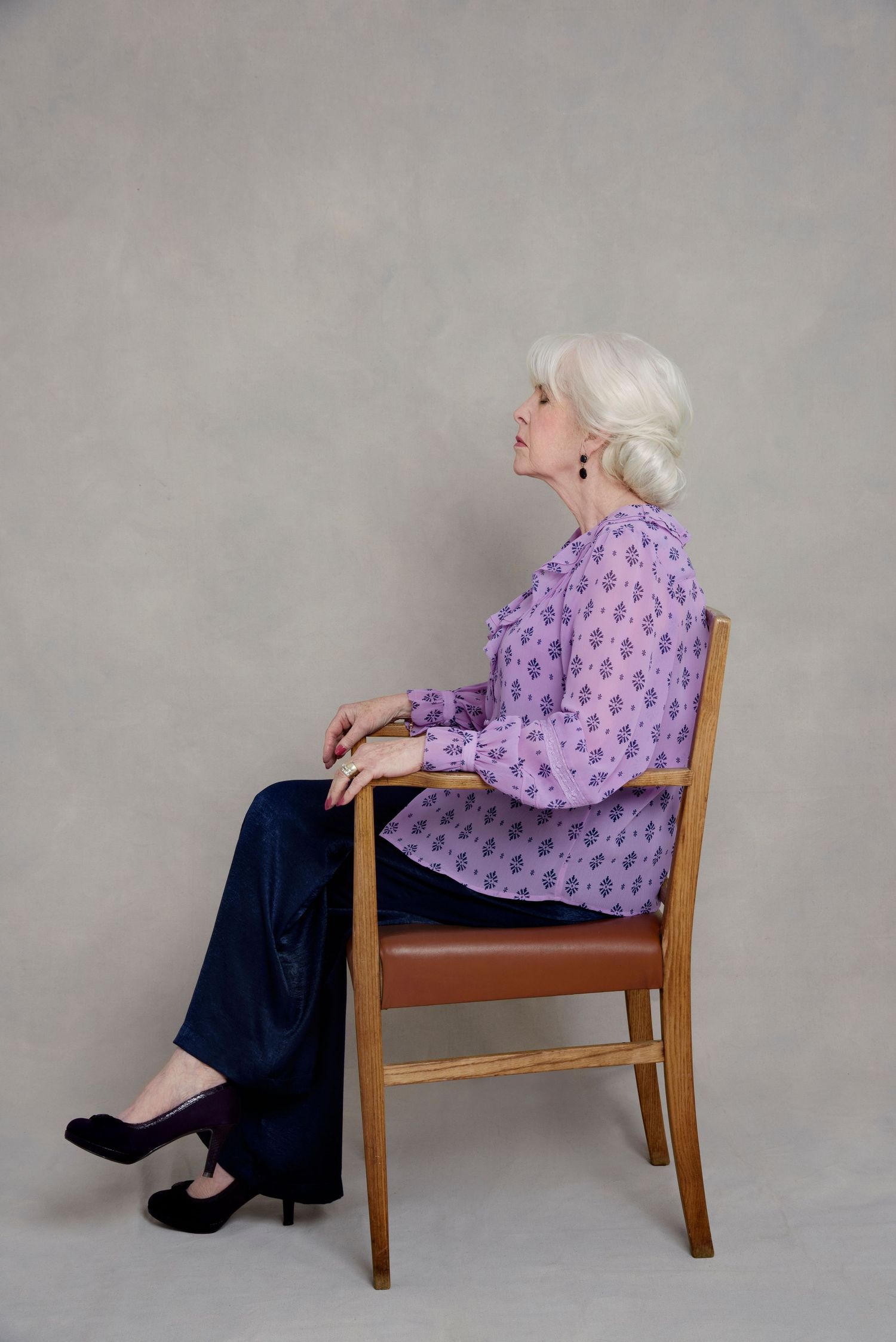 Person in a purple floral blouse and navy pants sits elegantly in a wooden chair against a neutral backdrop.
