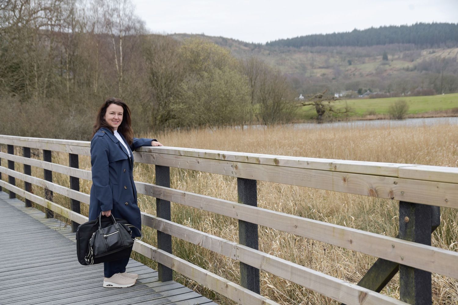 Woman in navy coat stands on wooden bridge overlooking reed beds and green hills in rural landscape.