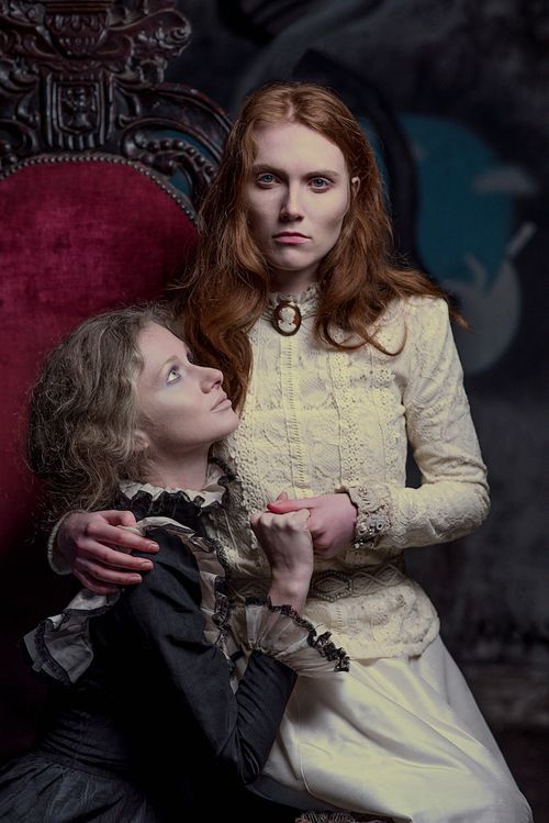 Two women in Victorian-era costumes pose dramatically, one seated in an ornate chair holding the other's hands.