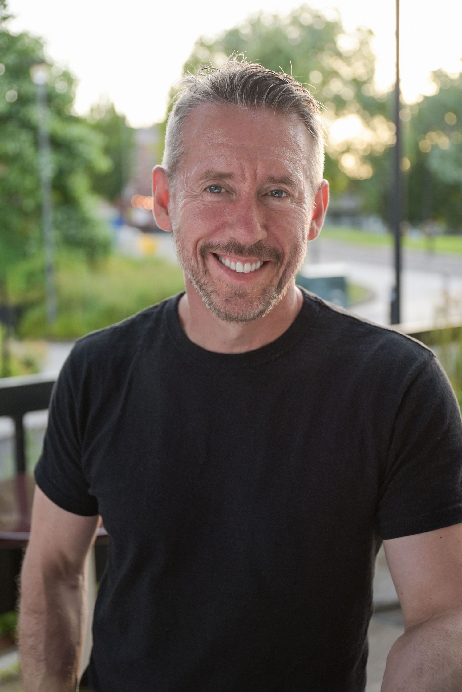 Smiling middle-aged man with gray hair and beard wearing a black t-shirt outdoors in a park setting.