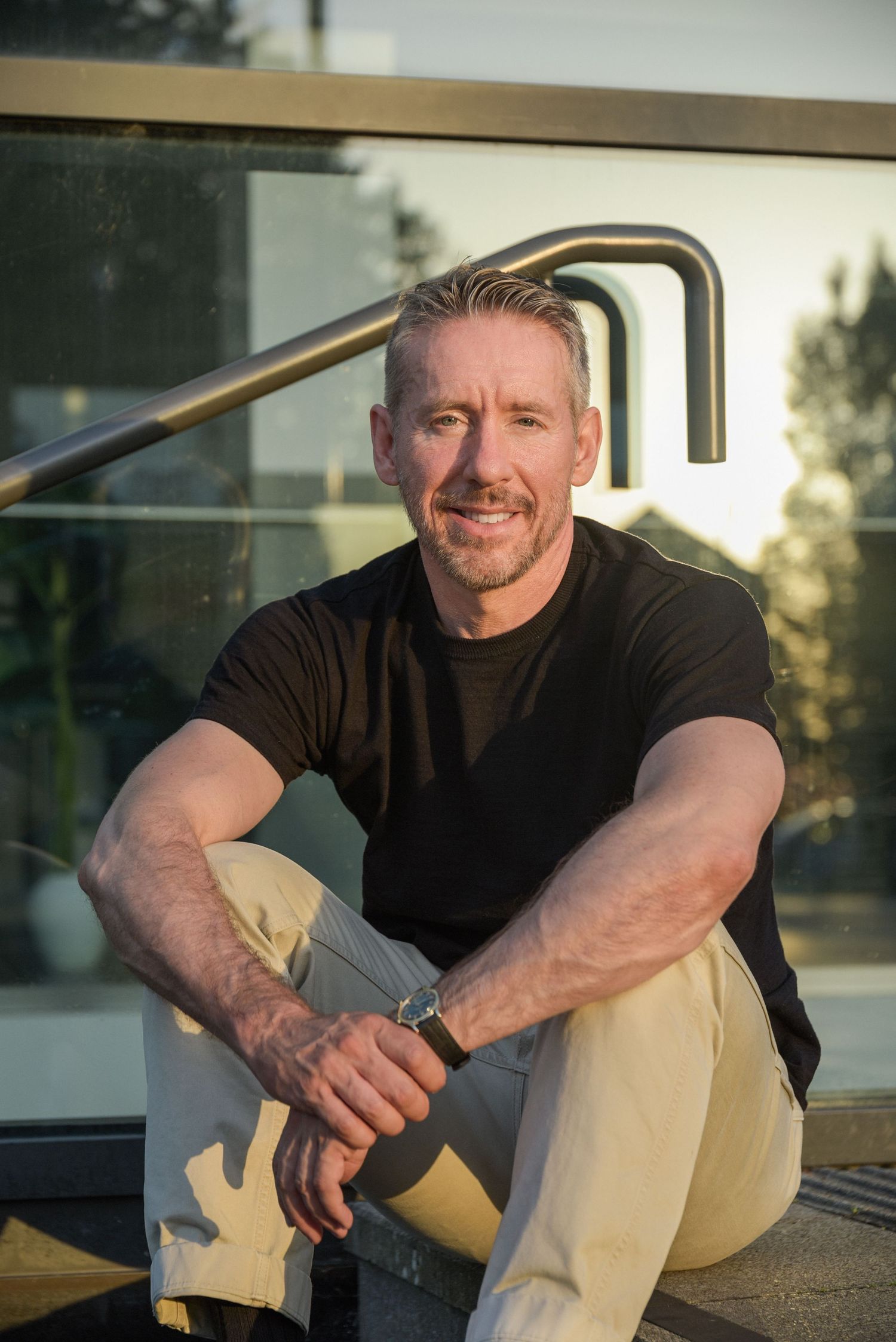 Middle-aged man with gray hair smiling, sitting on outdoor steps in a black t-shirt and khaki pants.