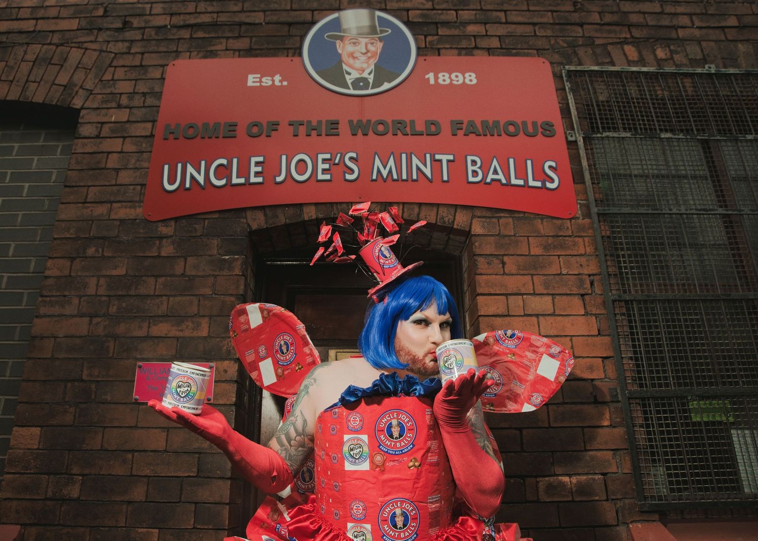 Person in red costume poses outside Uncle Joe's Mint Balls factory, holding candy tins, wearing blue wig.