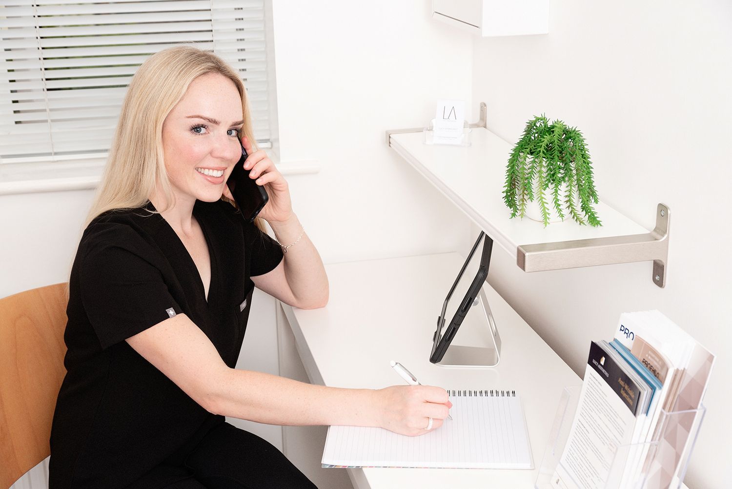 Smiling blonde receptionist in black dress talking on phone while taking notes at a modern office desk.