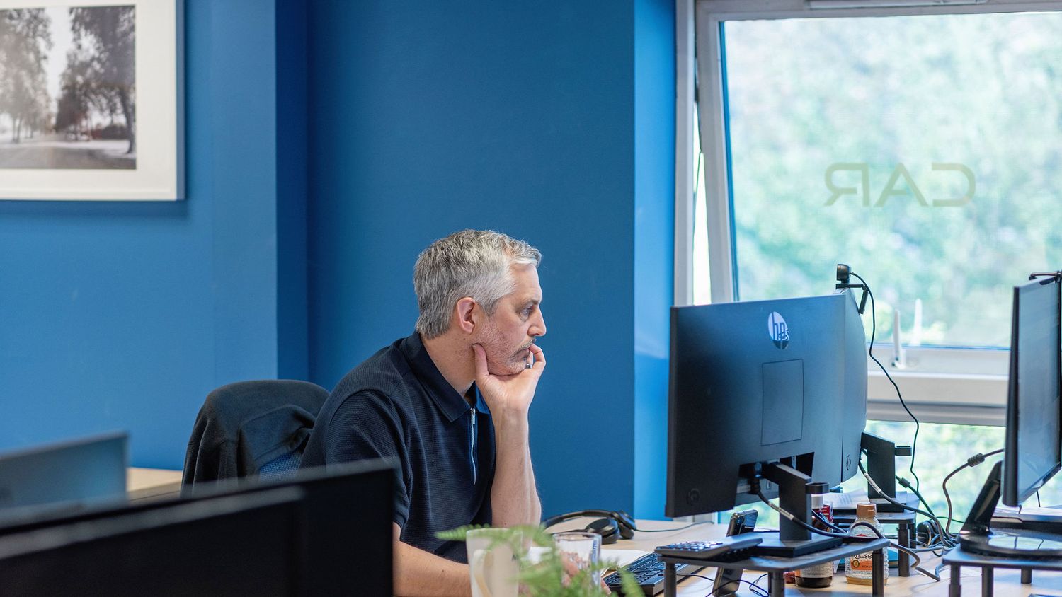 Professional man with gray hair sitting at desk with multiple monitors in a modern blue office workspace.