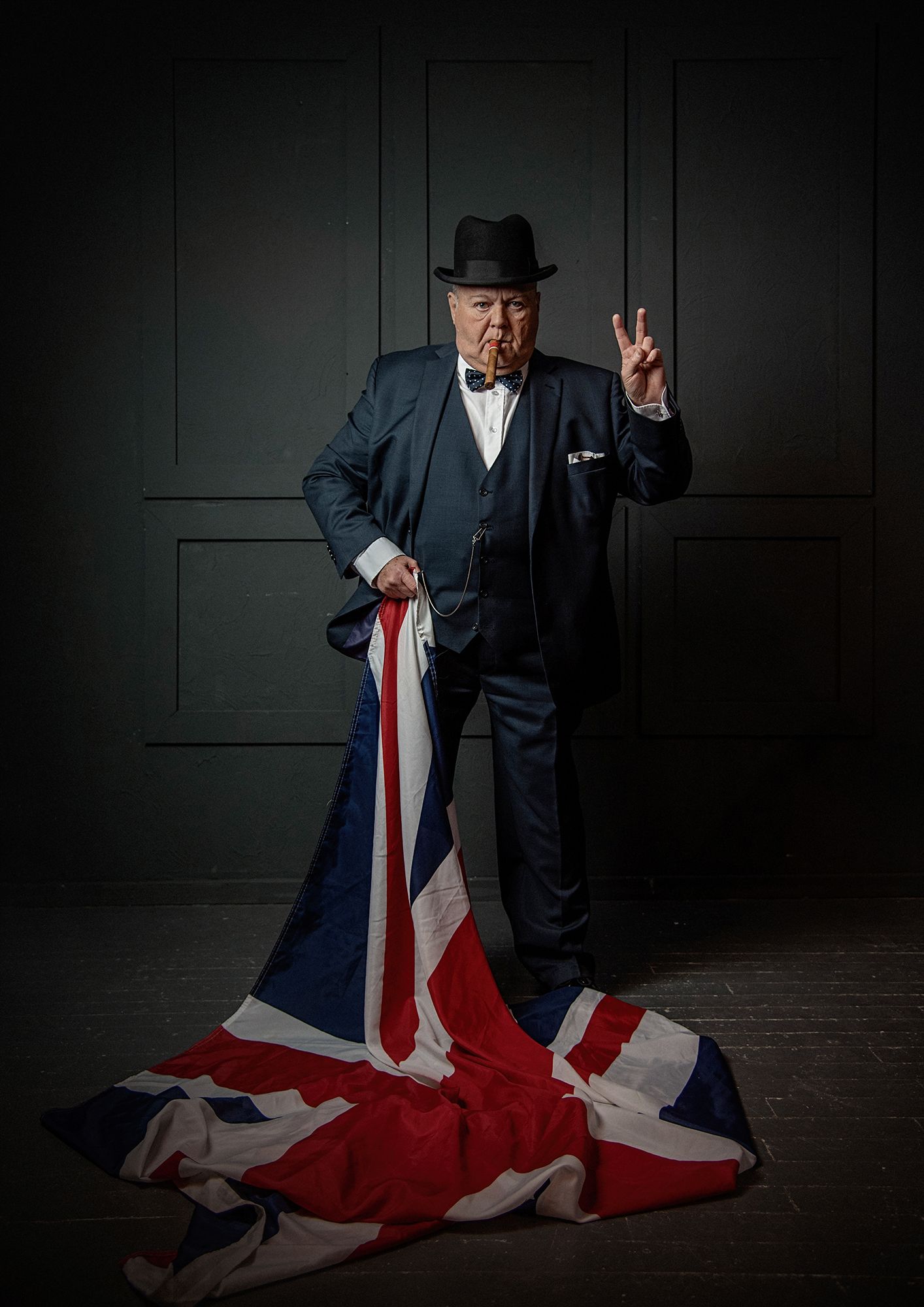 Figure in suit standing on Union Jack flag making peace sign against dark background.