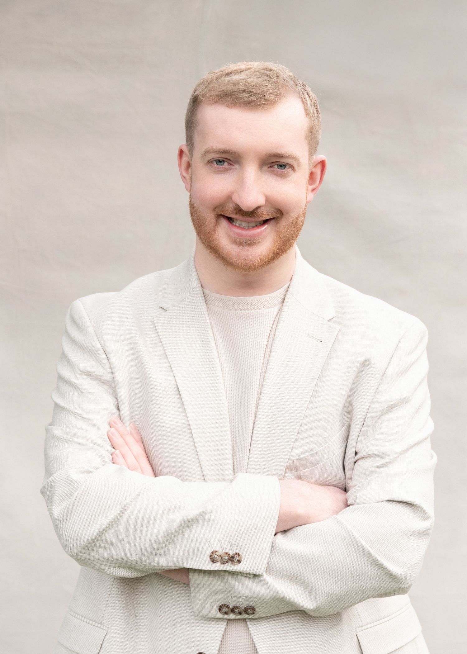 A person wearing a white formal suit stands with a confident pose and folded arms against a light background.