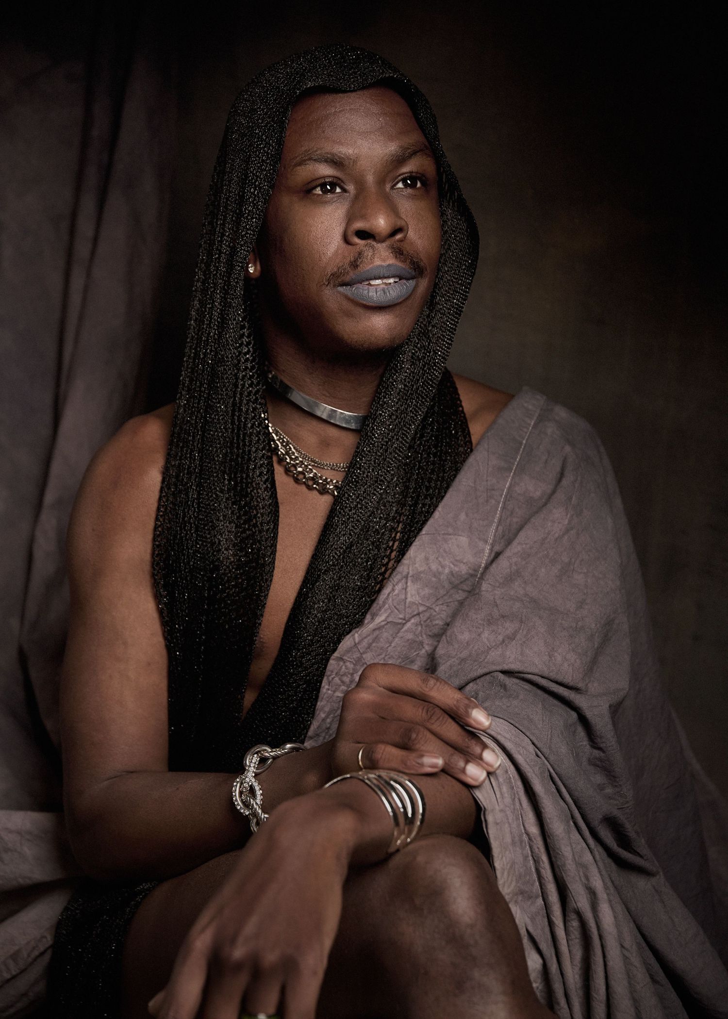 Artistic portrait featuring long braids, jewelry and draped fabric in moody lighting.