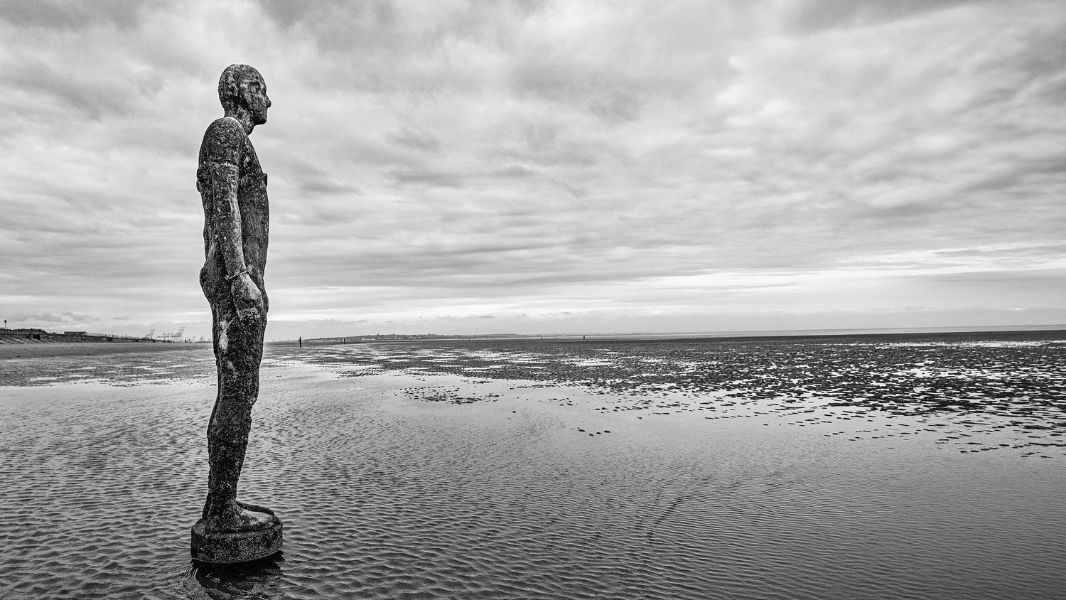 Black and white photo of an Iron Man statue on a reflective beach under a cloudy sky.