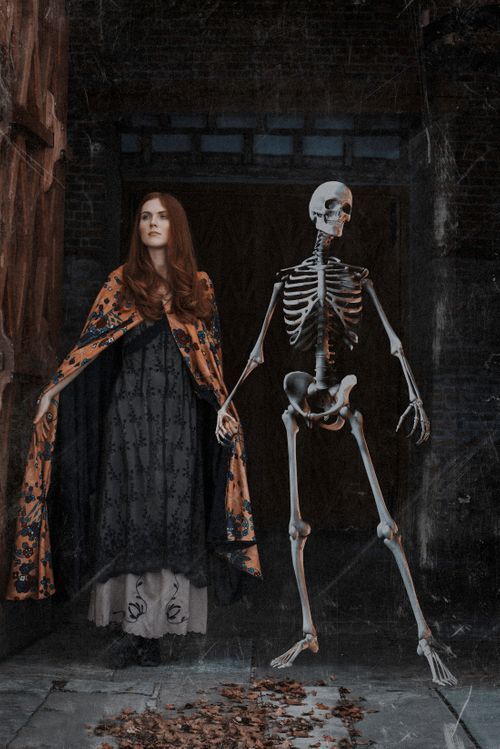 A red-haired woman in a gothic black gown and orange floral cape stands beside a full-size skeleton in a dark doorway.
