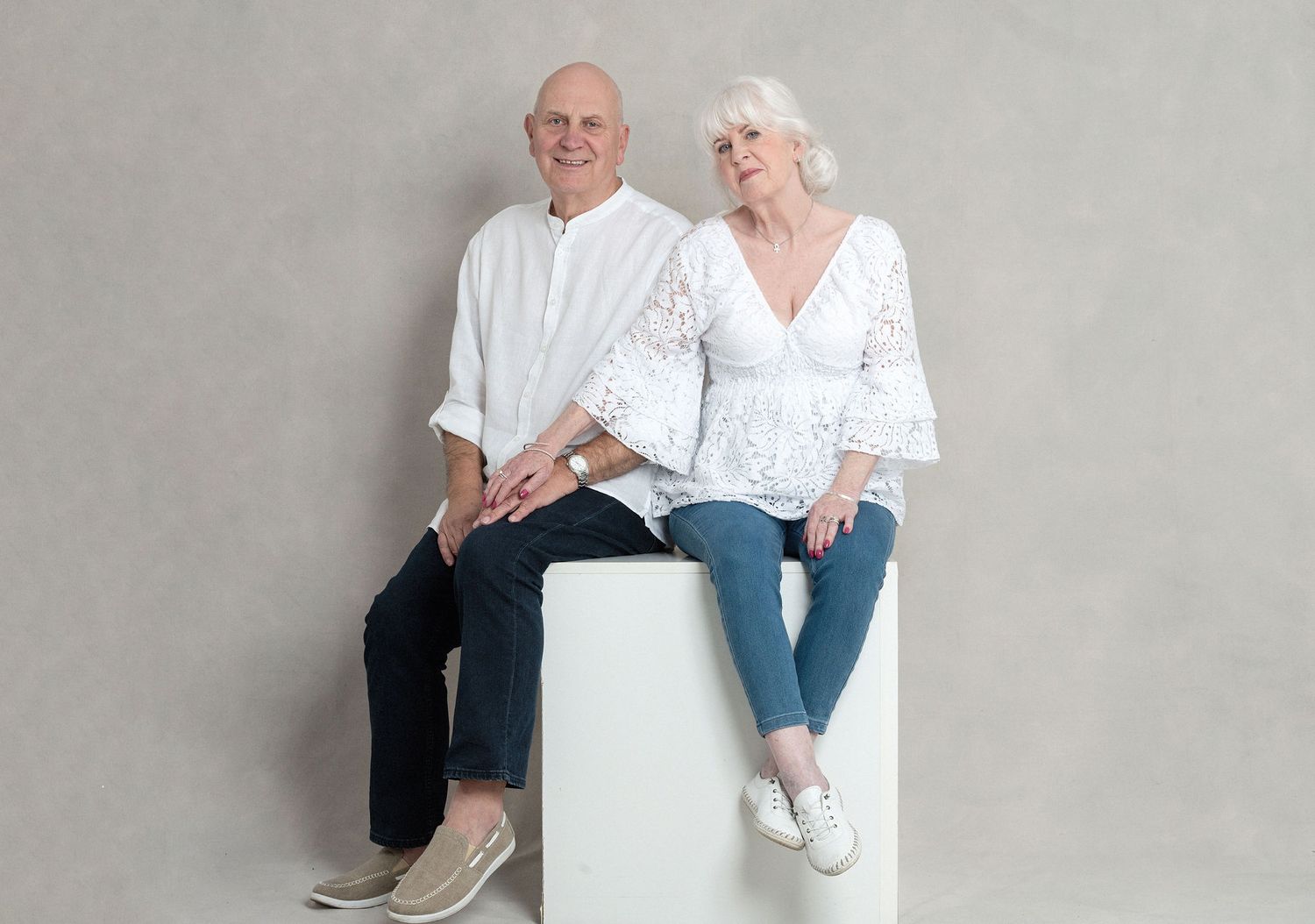 Couple in white tops and dark pants pose casually on a white cylindrical pedestal.