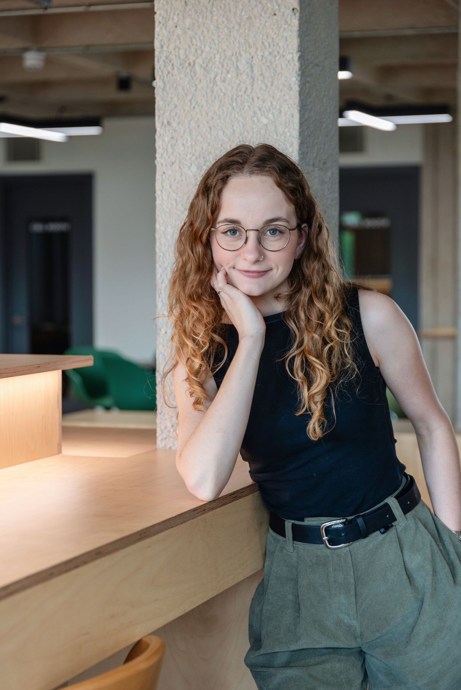 Young woman with curly red hair and glasses leaning on wooden counter in modern office space.