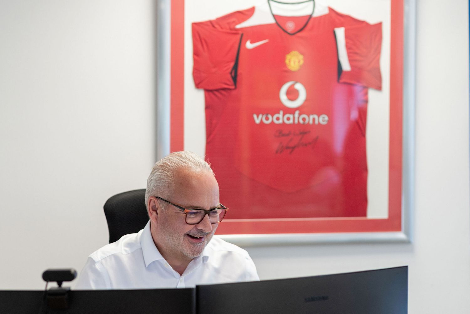 Smiling man at desk in office with framed signed Manchester United Vodafone red jersey displayed on wall behind him.