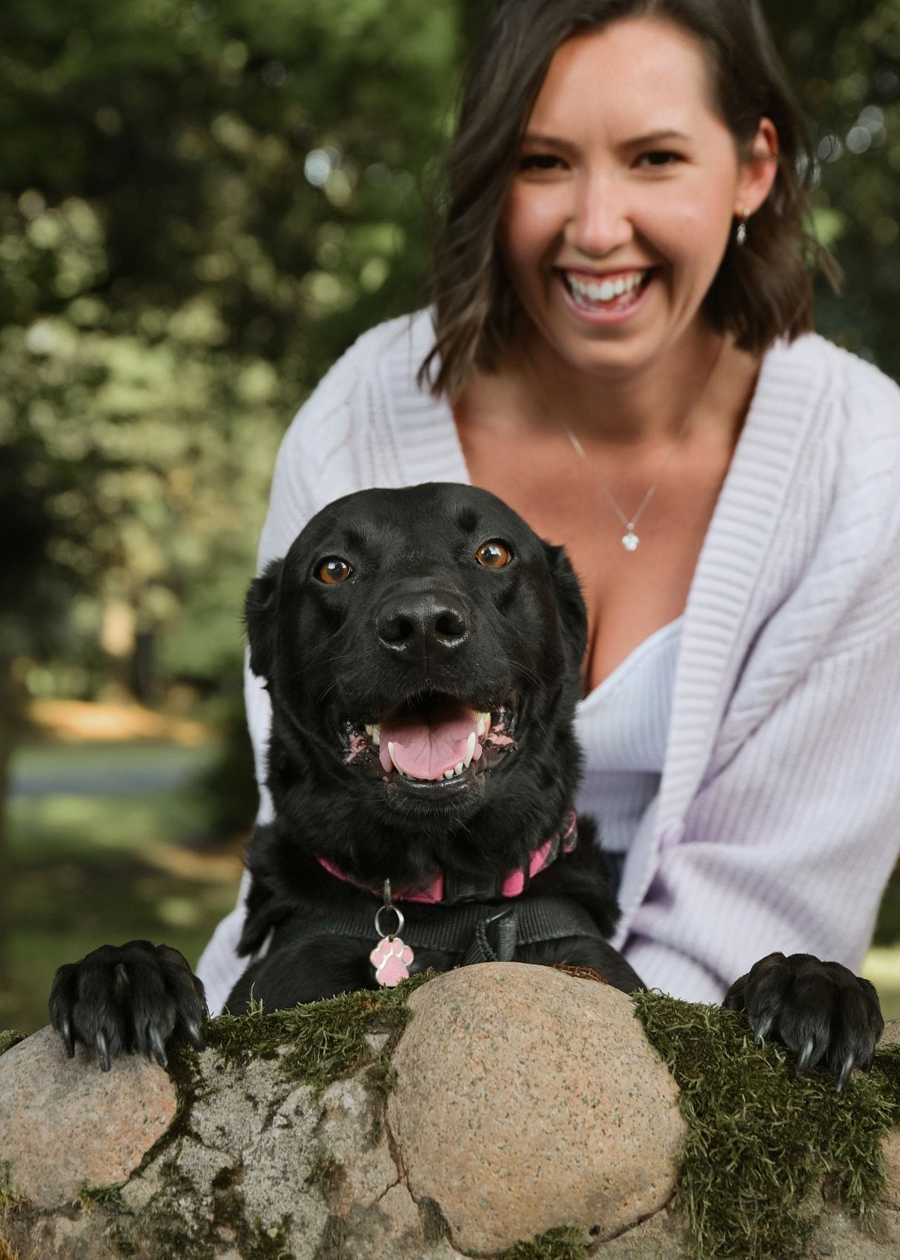 Smiling woman with a happy black Labrador dog posing outdoors on a mossy stone wall in a lush green park.