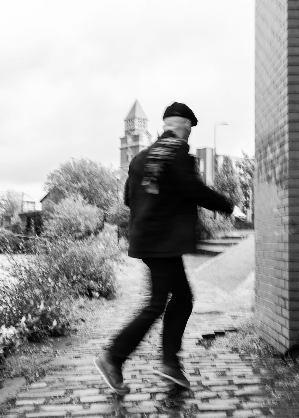 Black and white photo of a man in a beret dancing on cobblestones near a brick wall with a church tower behind.