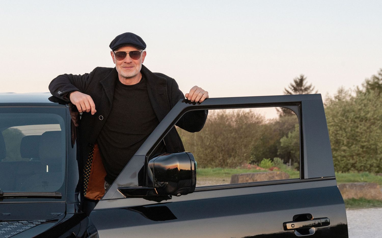 Man in black cap and sunglasses leaning on open door of black pickup truck outdoors at dusk.