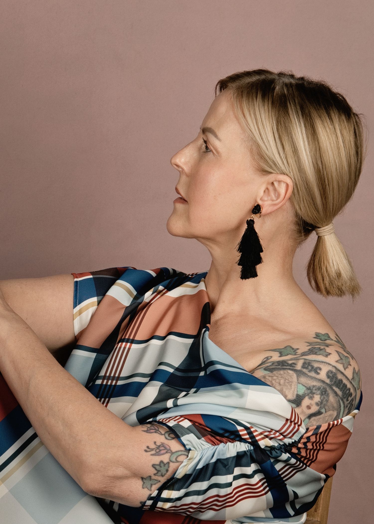 Blonde woman in striped off-shoulder top with tassel earrings and tattoos poses in profile against pink background.