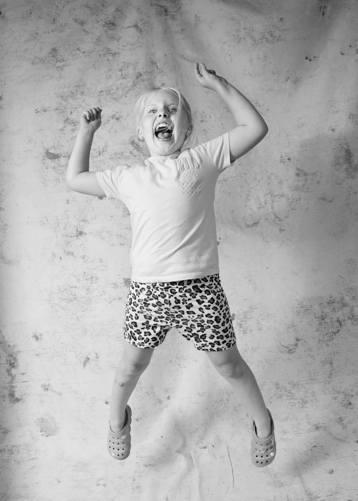 Black and white photo of an excited young child jumping with arms raised, wearing a white shirt and leopard print shorts.