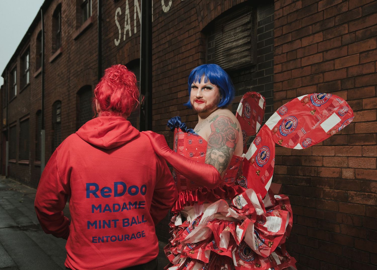 Person in red ReDoo hoodie poses with blue-haired performer in red costume near brick wall.