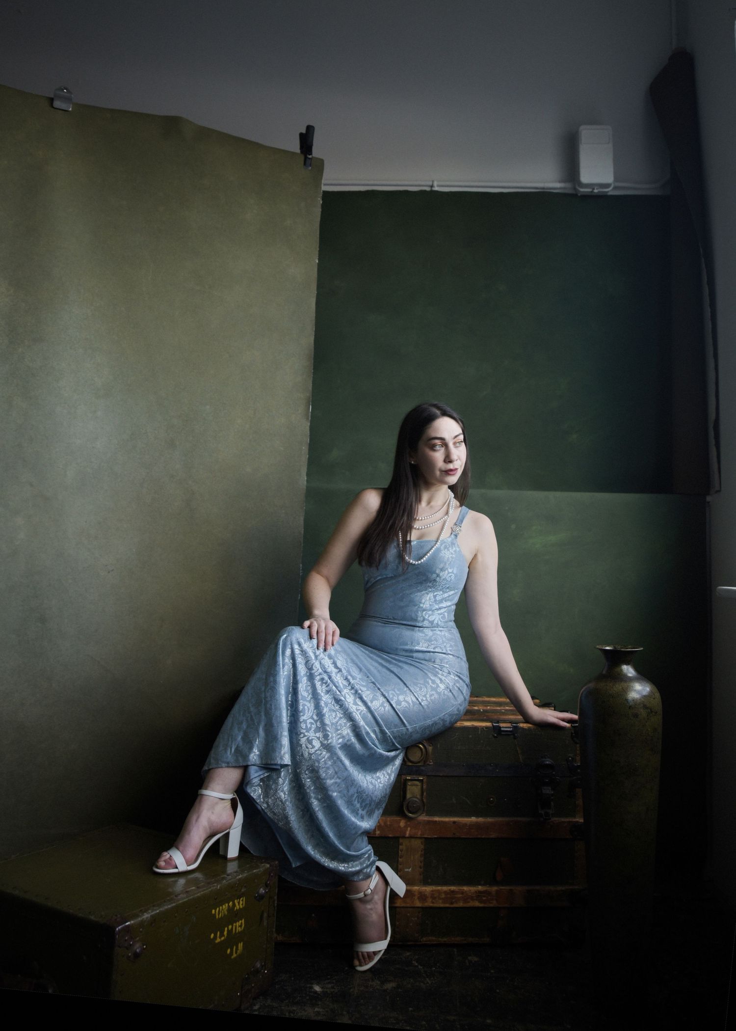 Vintage-inspired portrait of model in blue dress seated on wooden crate against dark green wall.