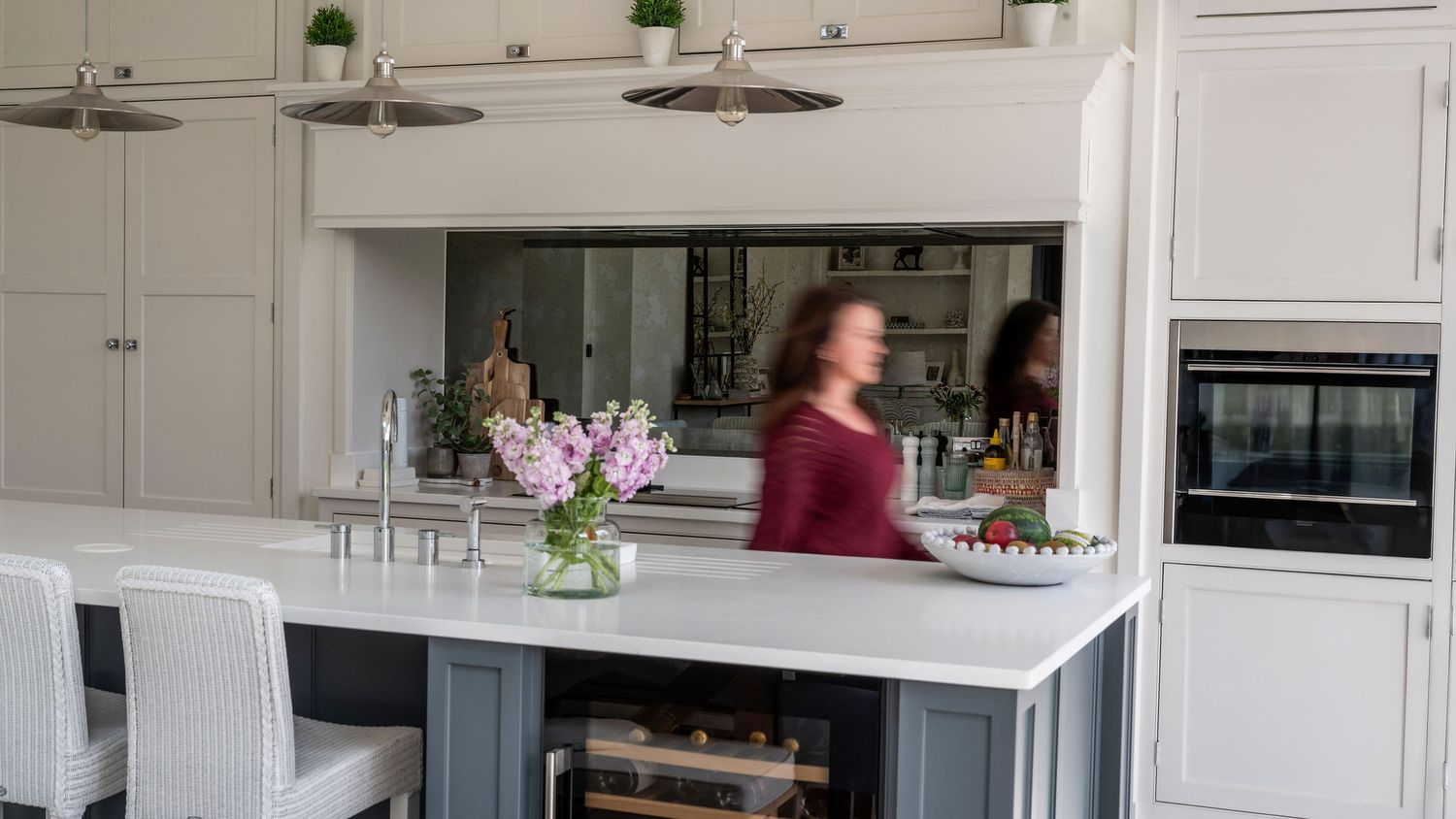 Woman in a modern white kitchen with gray island, marble countertops, mirrored backsplash, and pendant lights.
