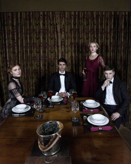 Elegantly dressed group at a formal dinner table with wine glasses and white plates in a dark, dramatic setting.