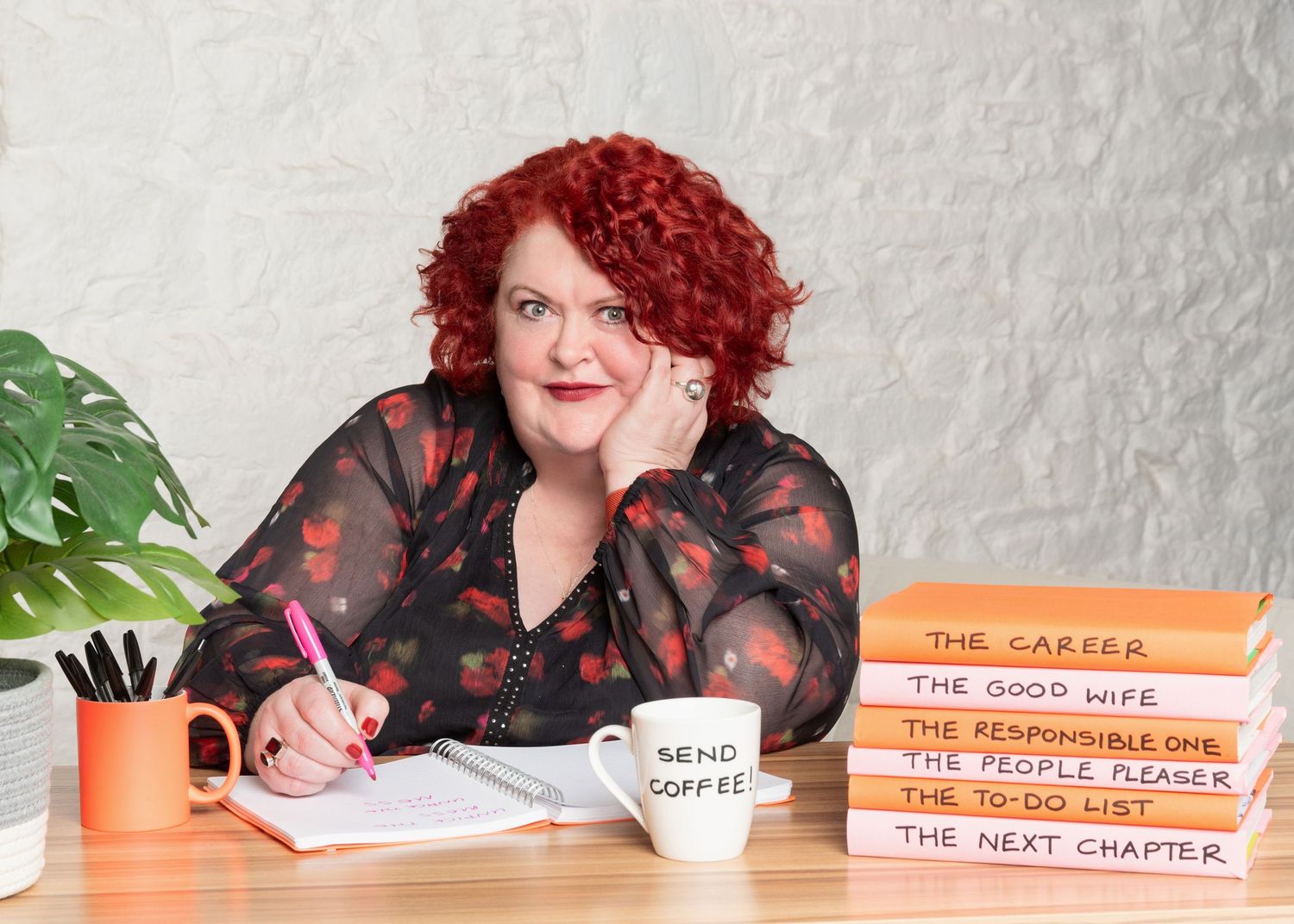 Woman with red curly hair and floral blouse rests her chin on her hand at a desk with a pen, notebook, 'Send Coffee' mug and stacked books.