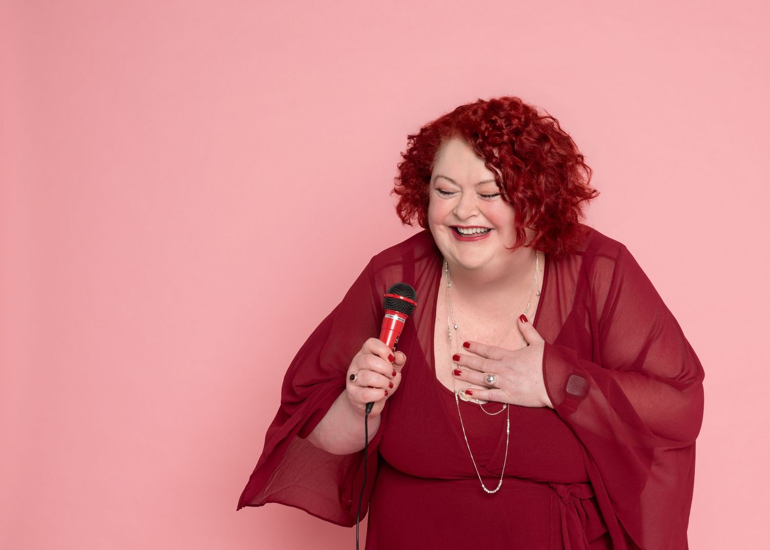 Smiling woman with curly red hair in a burgundy top laughs while holding a red microphone against a pink background.