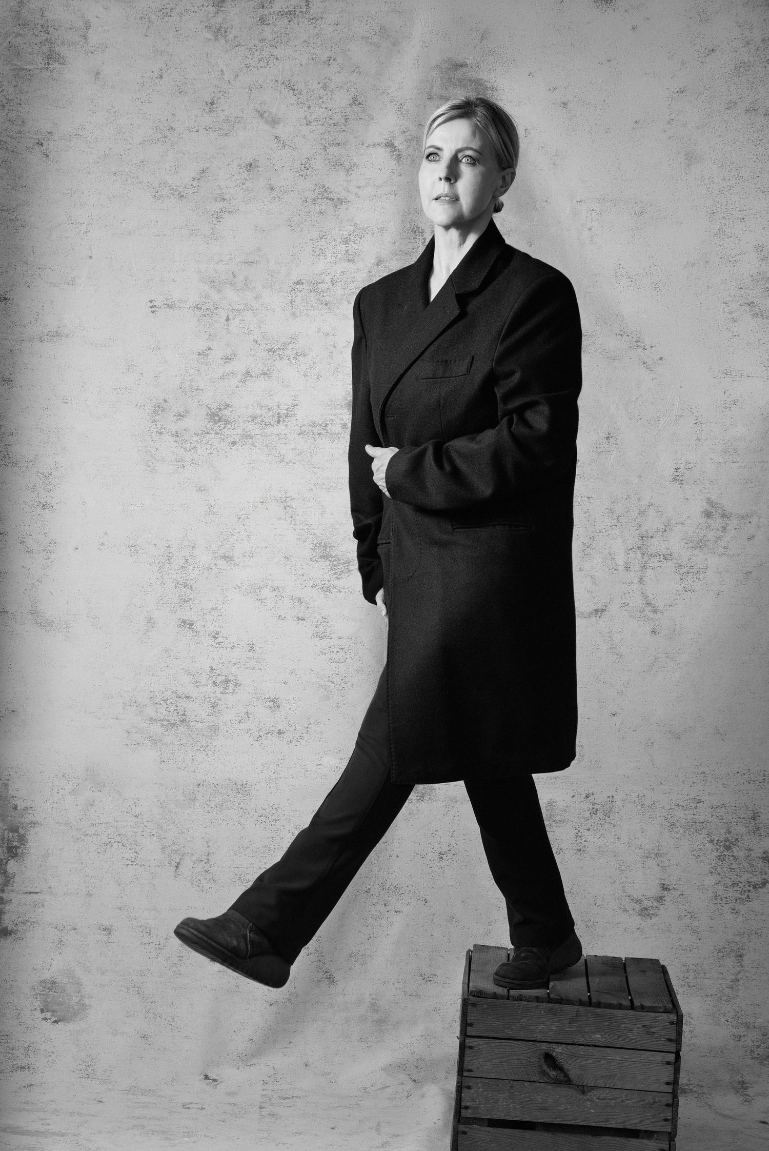 Black and white portrait of a woman in a dark oversized coat standing on a wooden crate against a textured wall.
