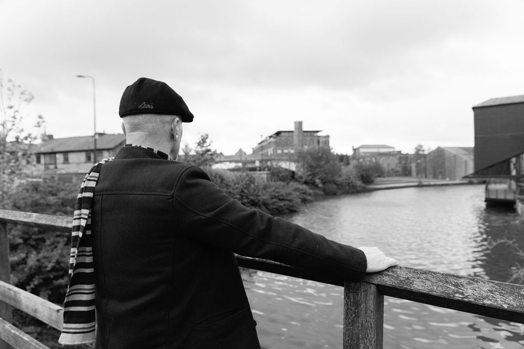 Elderly man in black jacket and beret leans on railing overlooking a canal in a black and white photo.