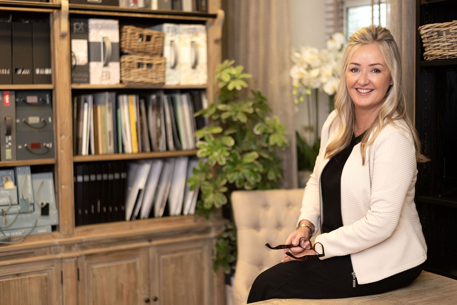 Business professional in white blazer seated in modern office with organized shelving and decor.