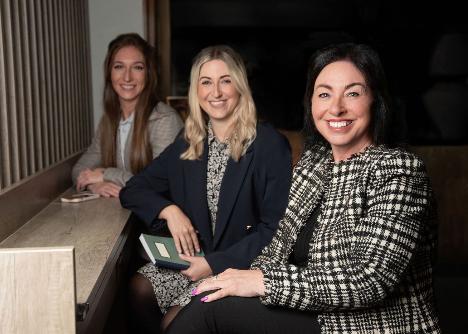 Three colleagues sit together on a bench in a hallway, wearing business attire and smiling.
