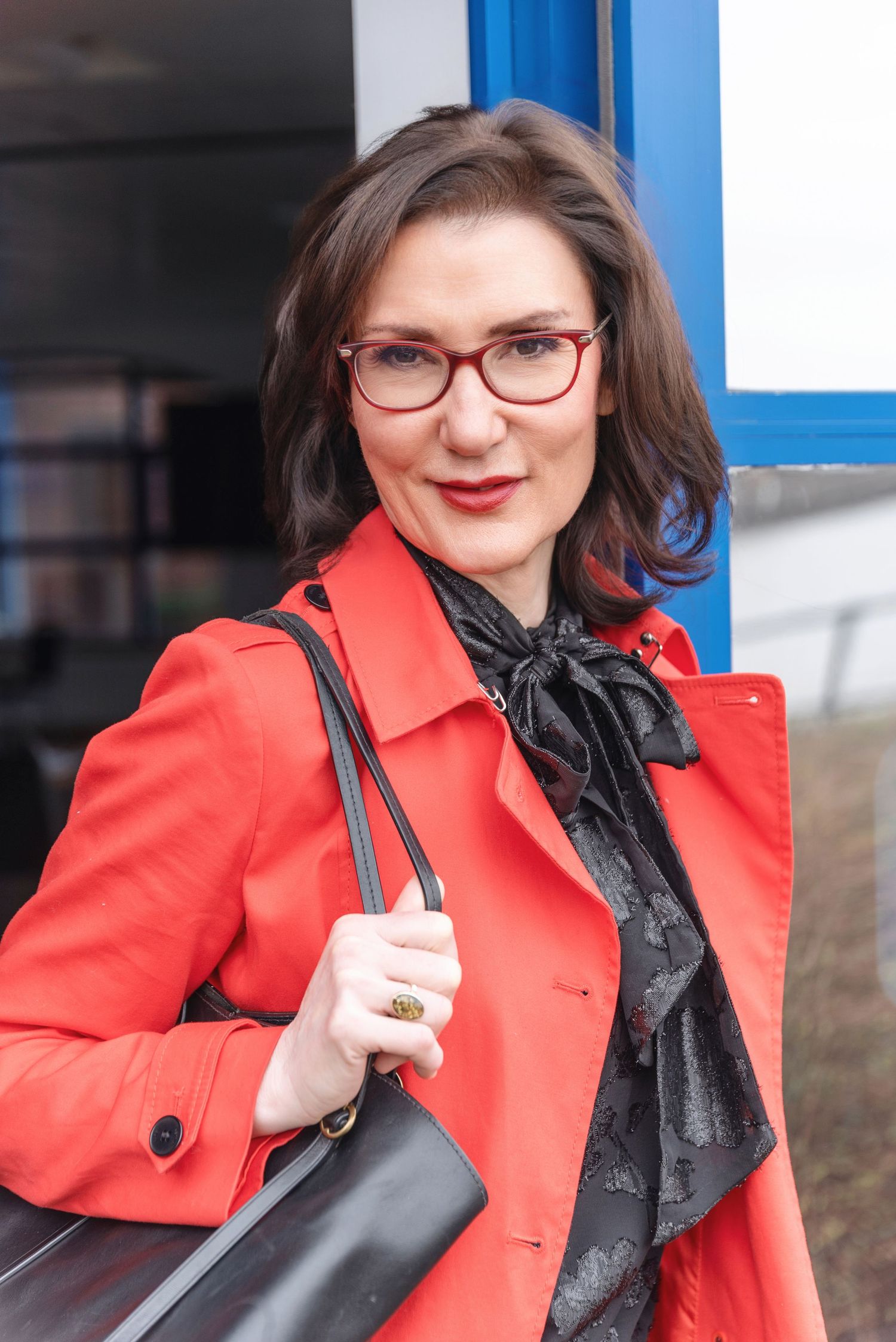 Professional portrait of someone wearing a vibrant red coat, glasses, and black scarf while carrying a shoulder bag.