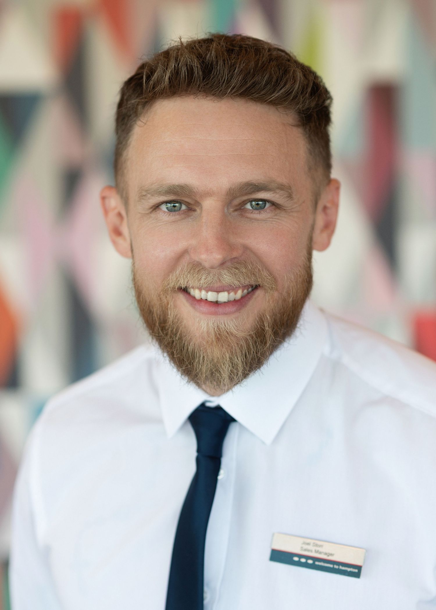 Professional headshot of someone wearing a white dress shirt, navy blue tie and name badge against a colourful blurred background.