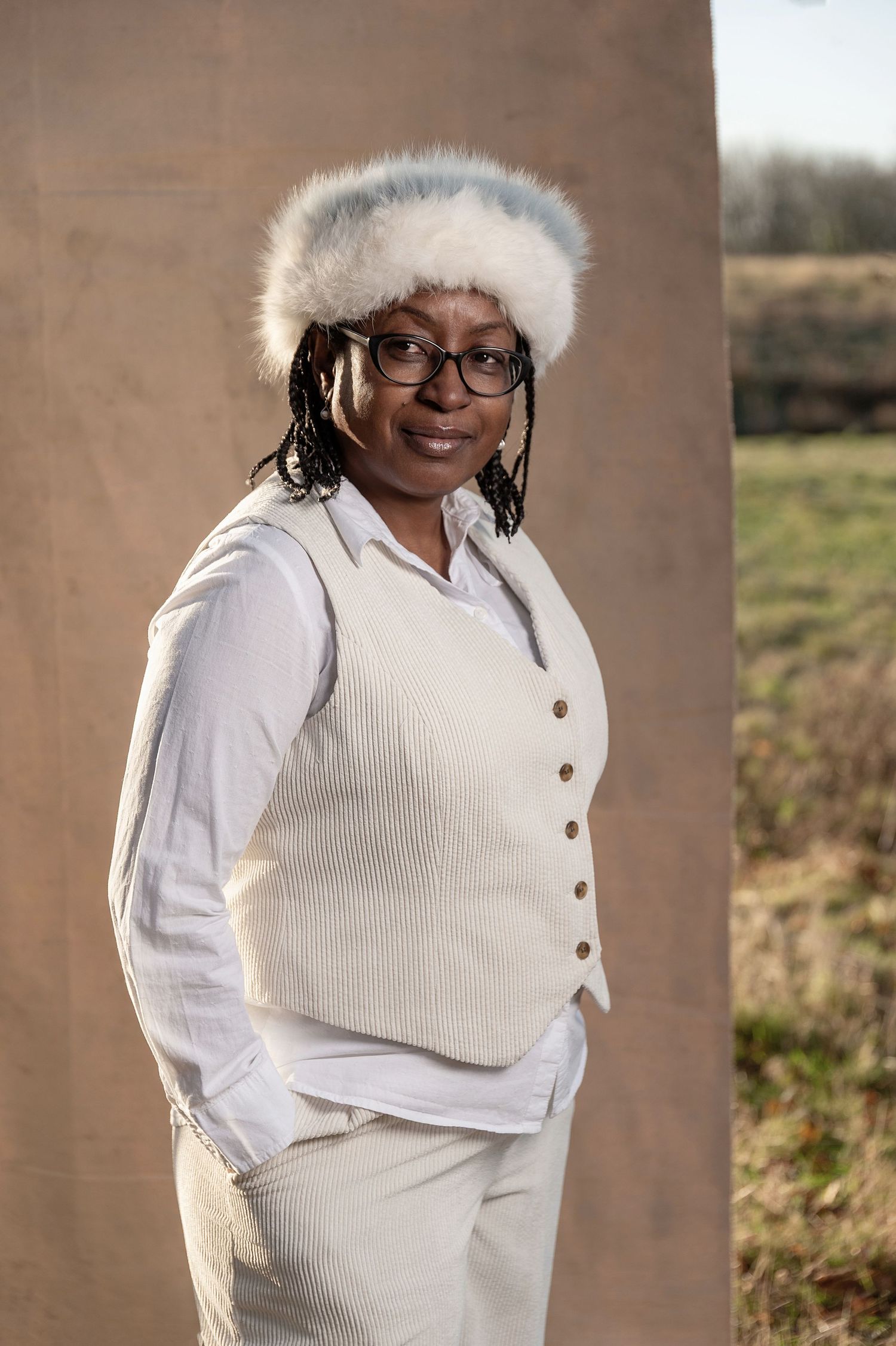 Person wearing glasses, white knit vest, button-down shirt and furry winter hat poses outdoors against brown wall.