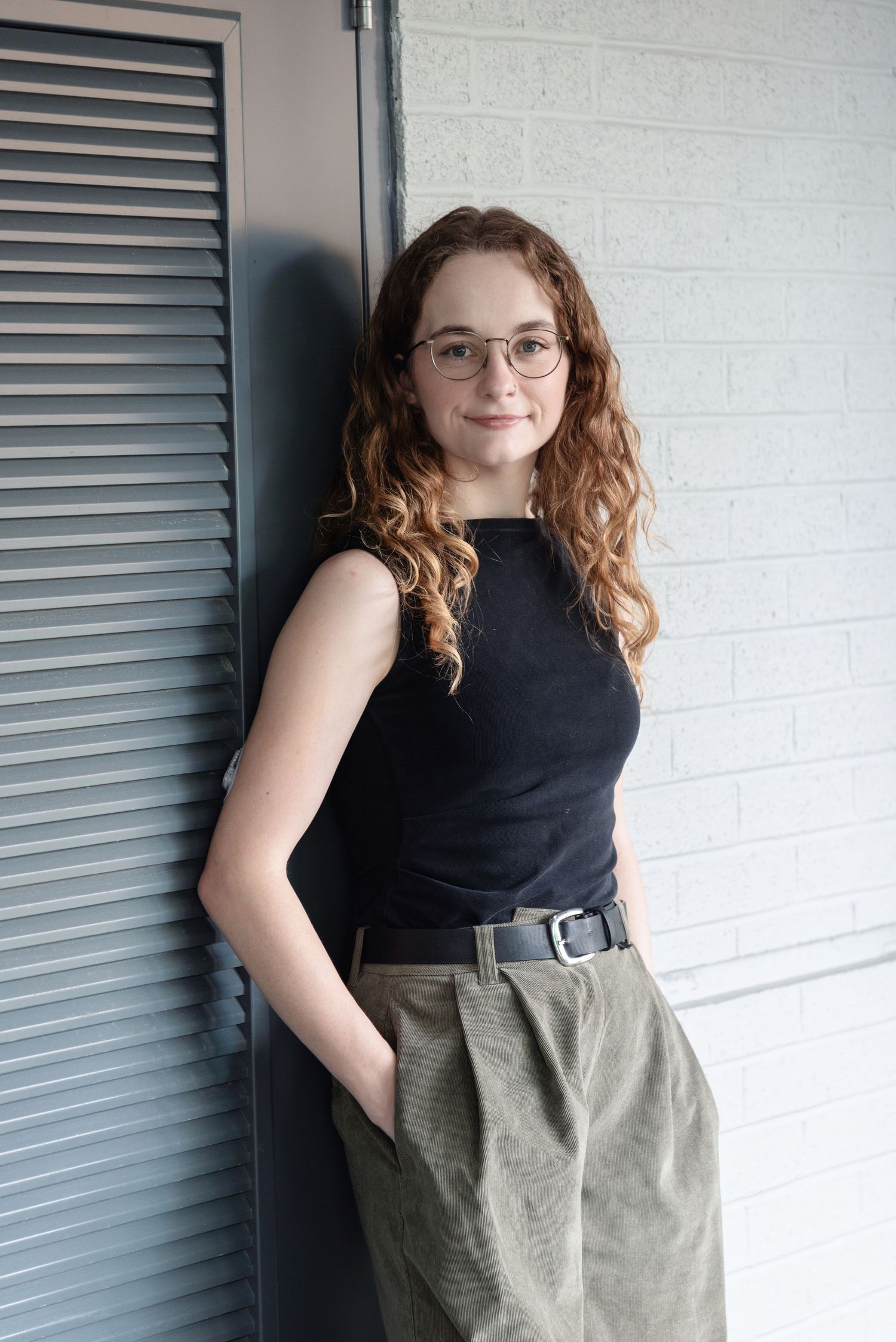Young woman with curly red hair and glasses wearing black top and olive trousers leaning against white brick wall.