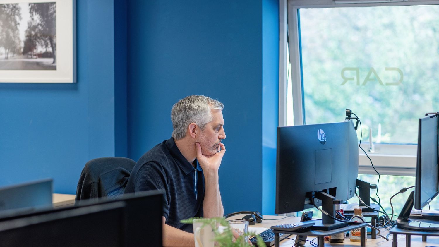 Man with gray hair sits at desk working on computer monitor in modern blue office workspace.