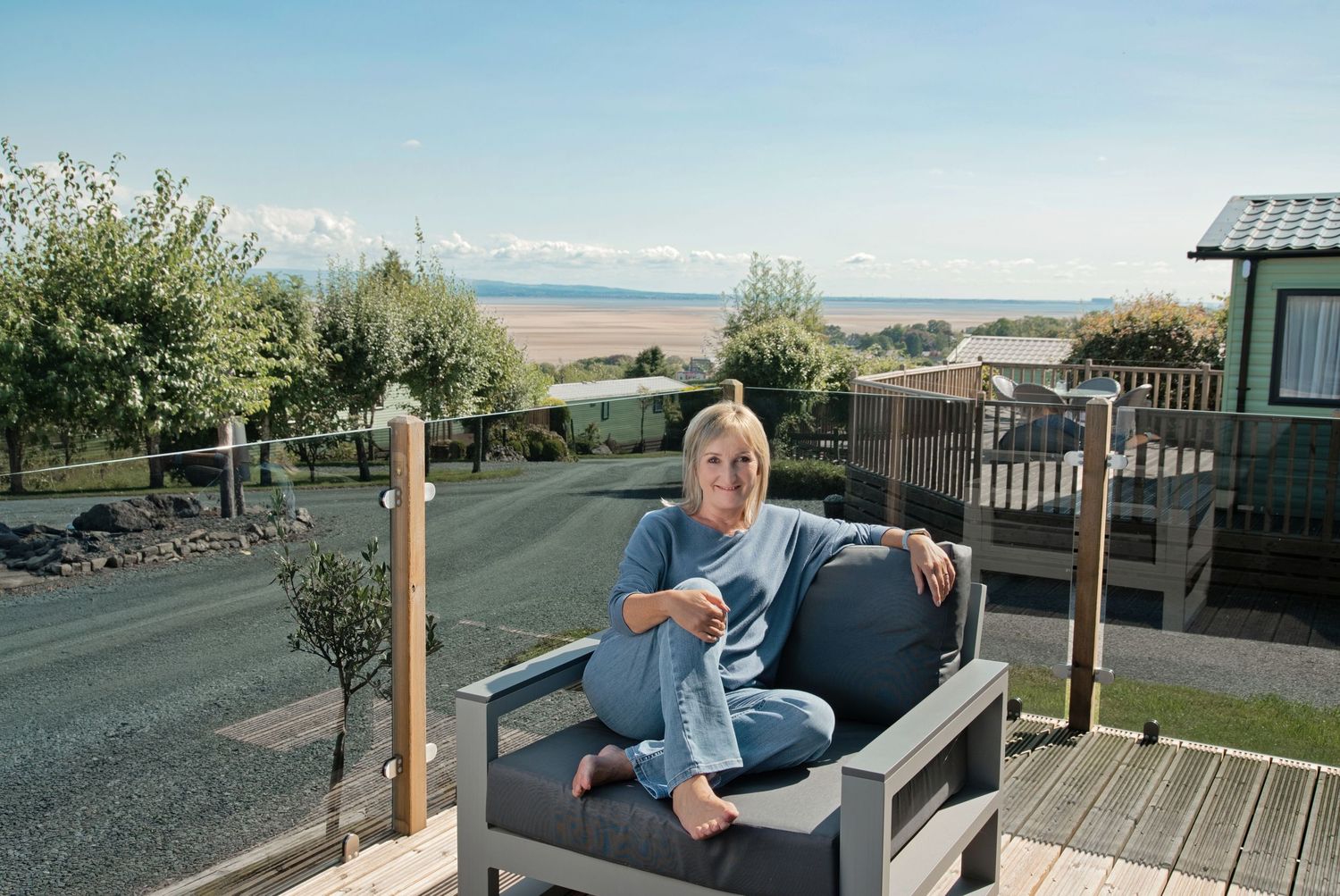 Woman relaxing on outdoor chair on holiday park decking with scenic coastal views and sandy beach in background.