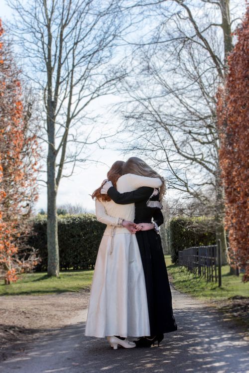 Two women in elegant dresses embracing on a tree-lined park pathway.
