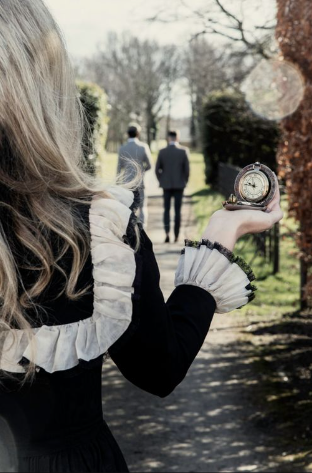 Woman in black ruffled dress holds vintage pocket watch as two men walk away on a park path.