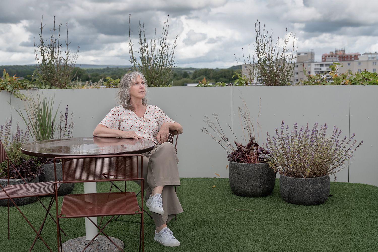 Older woman sitting alone at a metal patio table on a rooftop terrace with potted plants and cloudy sky.