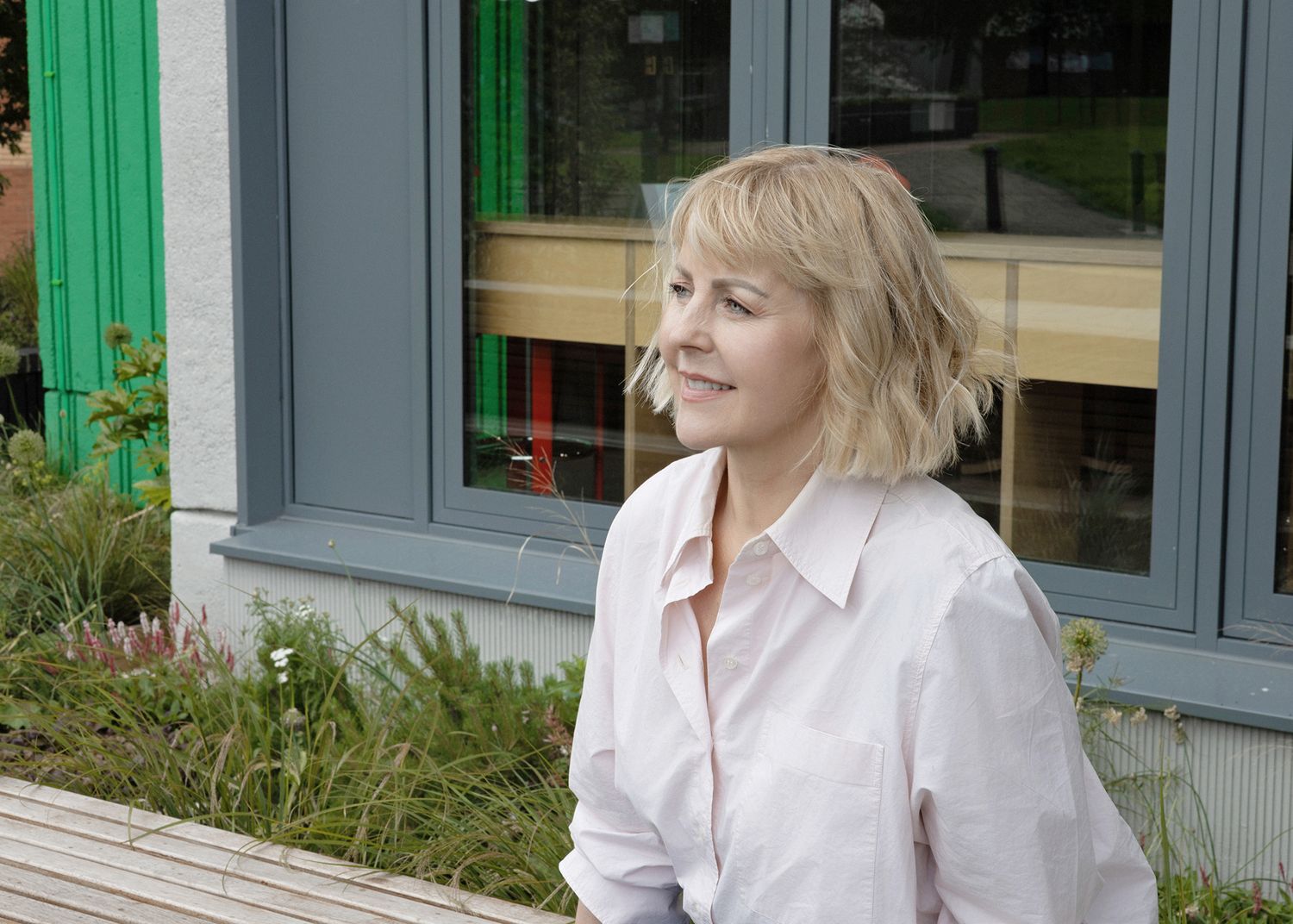Blonde woman in a pink shirt smiling outdoors near a modern building with green accents and lush plants.