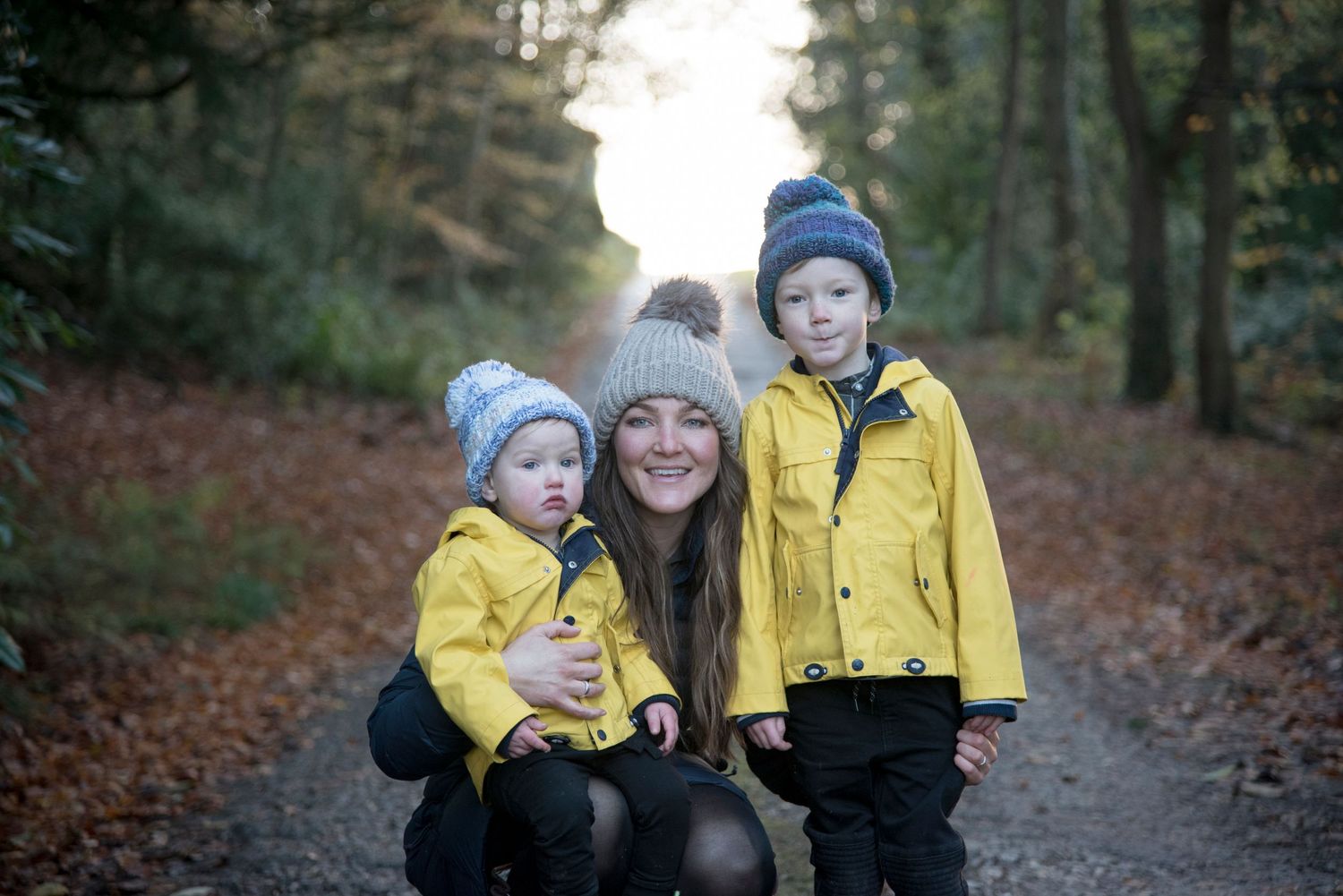 Family wearing yellow jackets and knit hats walks together on an autumn forest path surrounded by fallen leaves.