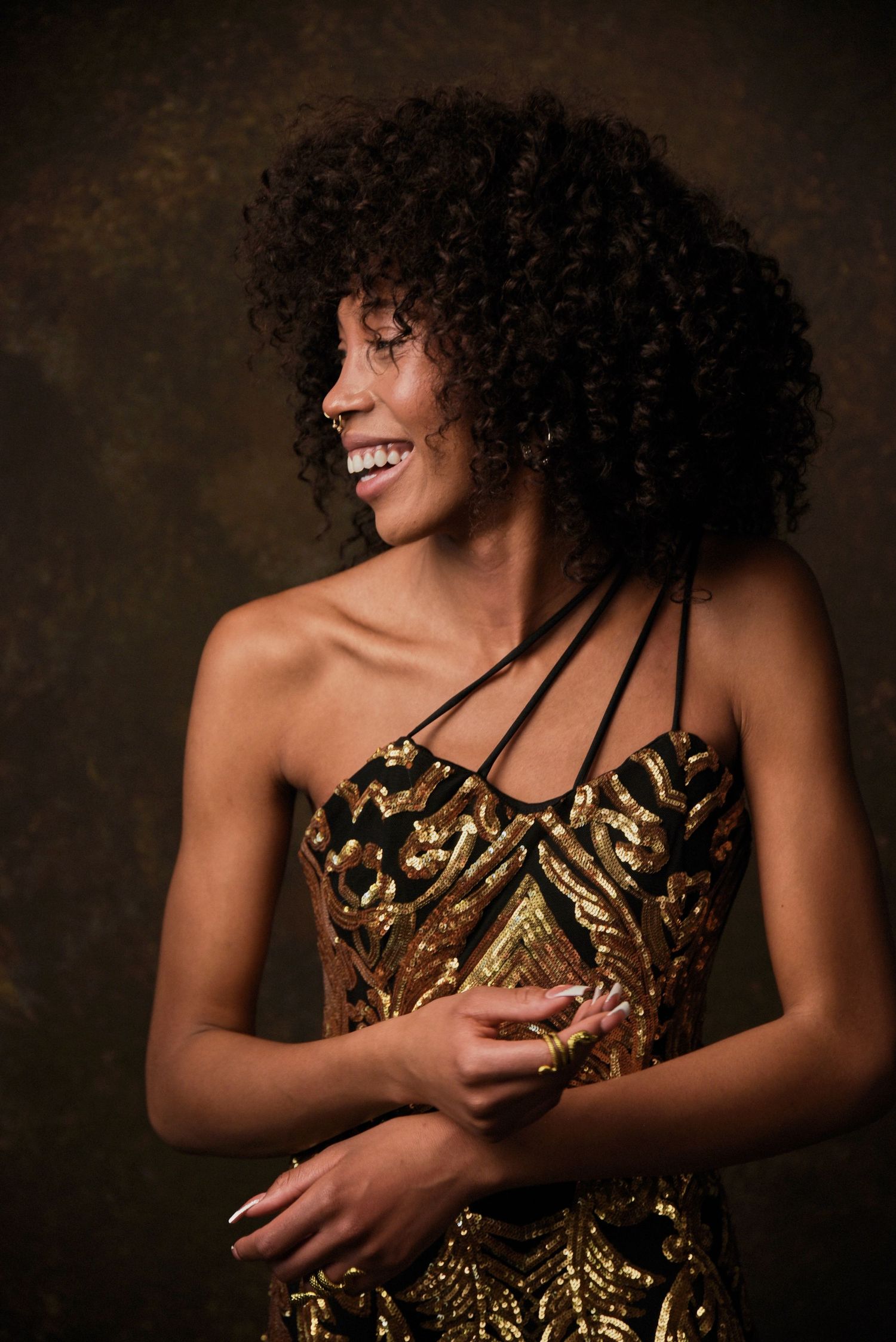 Portrait of someone in a glamorous gold sequined dress with natural curly hair against a dark background.